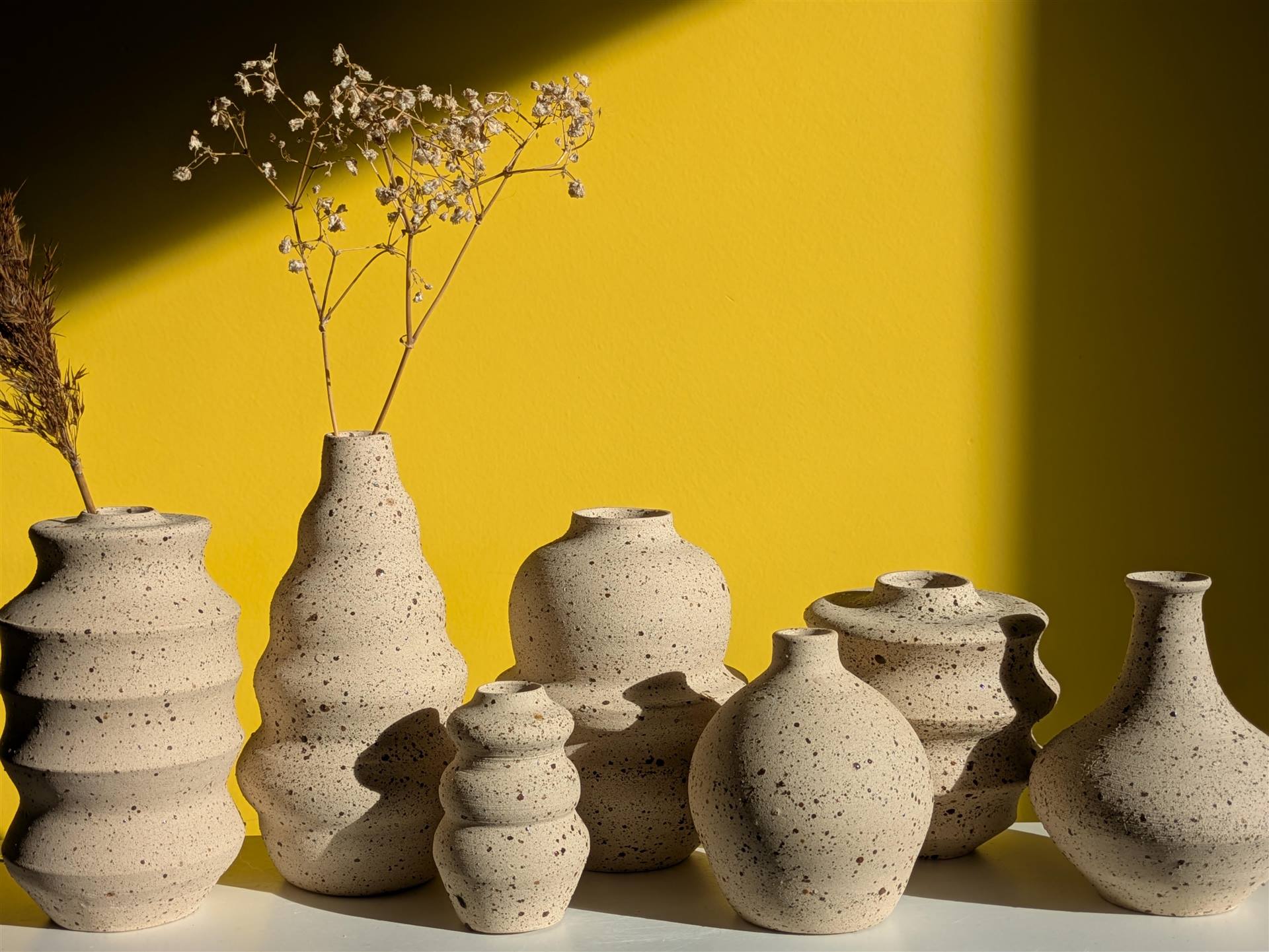 A row of handmade speckled ceramic vases in raking light against a yellow studio wall